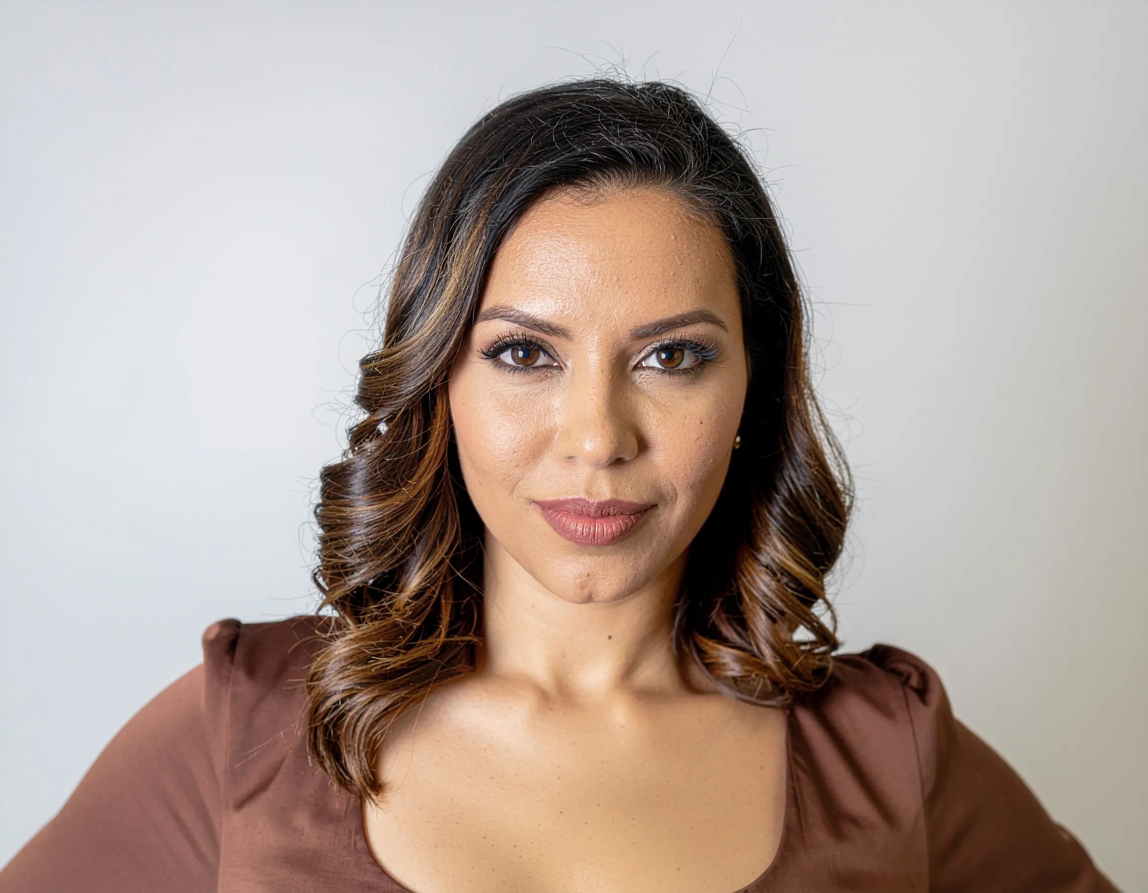 Close-up portrait of a woman with wavy highlighted brown hair wearing a brown top against a light gray background.