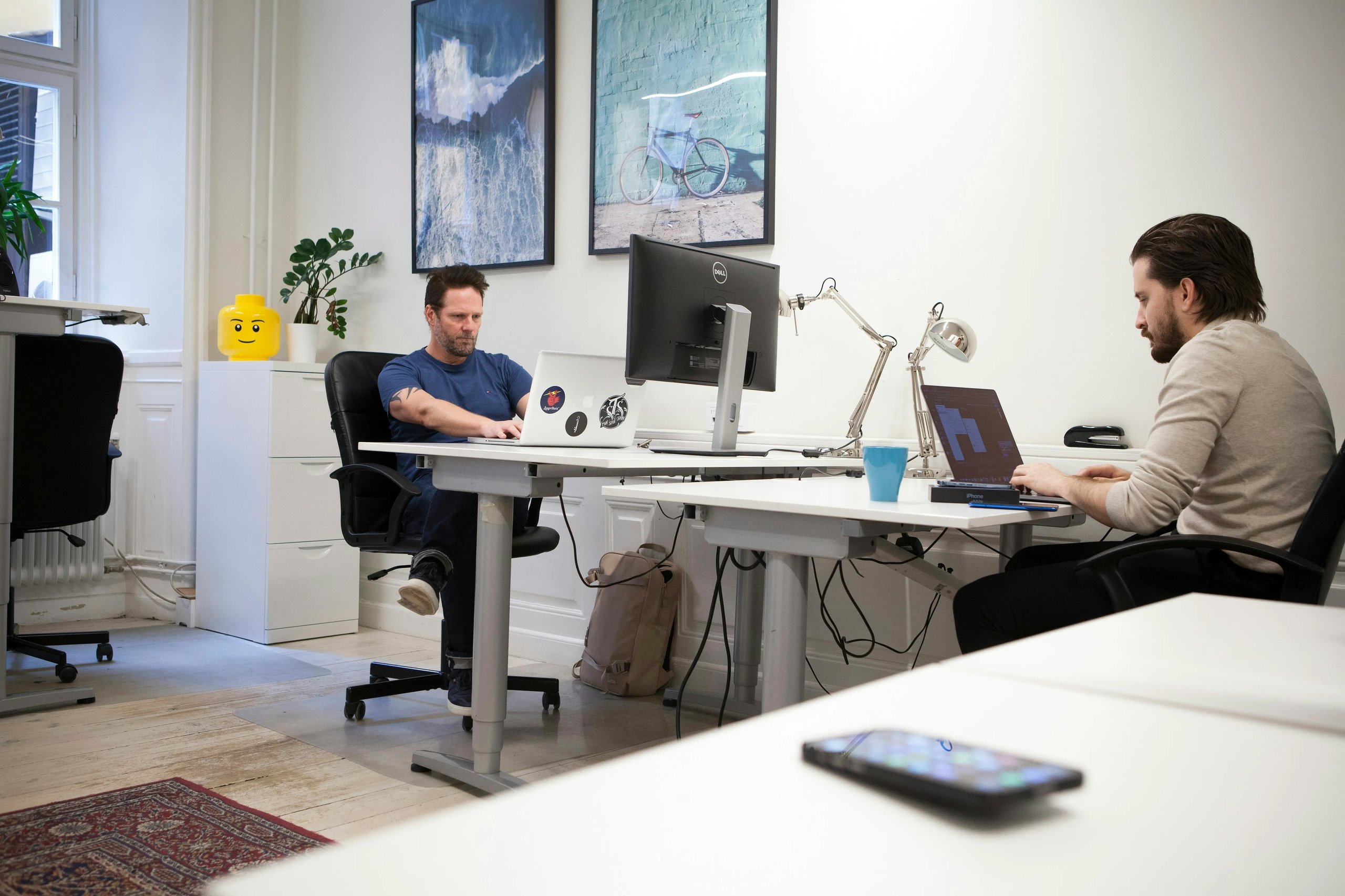 People work at desks with laptops and monitors in a bright, plant-filled home office.